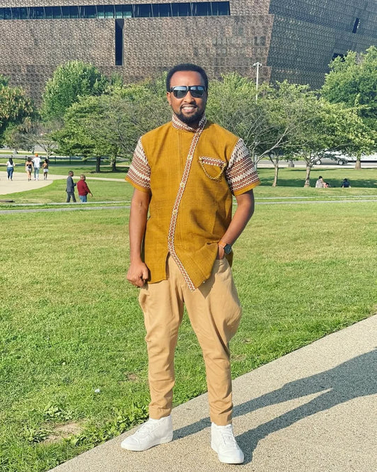Man in a mustard yellow shirt and beige pants standing on a sidewalk with a park in the background rown Modern Habesha Men’s Shirt Stylish Ethiopian Traditional Wear ethgebya gebeya usa habesha men