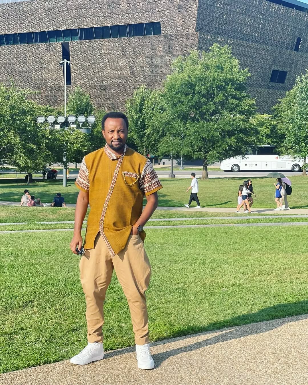 Man in a mustard shirt and beige pants standing on a grassy area with trees and a building in the background. rown Modern Habesha Men’s Shirt Stylish Ethiopian Traditional Wear ethgebya gebeya usa habesha men