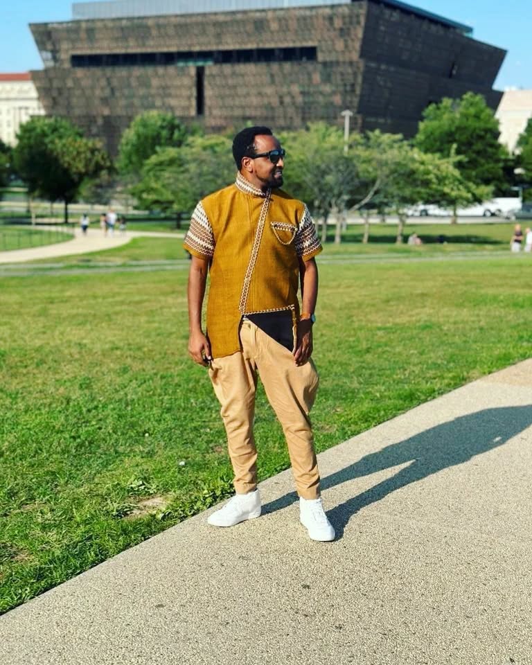 Man in mustard yellow shirt and beige pants standing on a sidewalk with a building and greenery in the background rown Modern Habesha Men’s Shirt Stylish Ethiopian Traditional Wear ethgebya gebeya usa habesha men