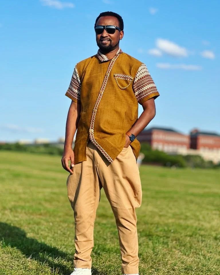 Man wearing a mustard-colored traditional outfit with sunglasses, standing in a grassy field. rown Modern Habesha Men’s Shirt Stylish Ethiopian Traditional Wear ethgebya gebeya usa habesha men