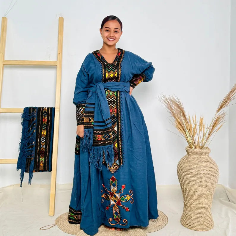 Woman wearing a blue embroidered dress standing in a room with a mirror and decorative vase. Dark Blue Raya Cultural Dress Handwoven Ethiopian Habesha Kemis ethgebya gebeya usa habesha kemis