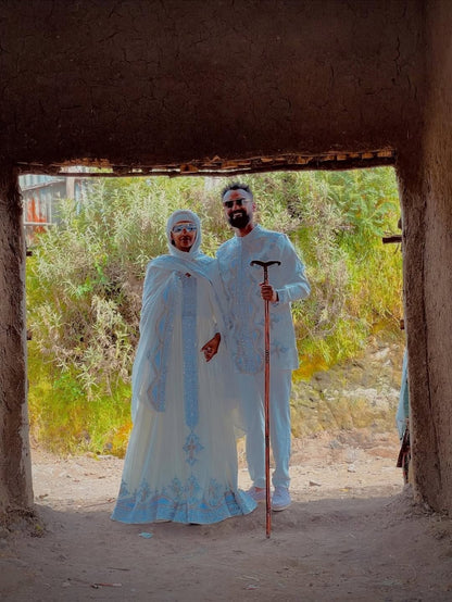 Two people in traditional attire standing in a desert setting with a natural archway. Silver Modern Habesha Couple Outfit Matching Ethiopian Attire ethgebya gebeya usa habesha kemis