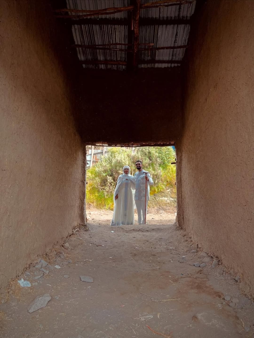 Two people in white clothing standing at the end of a narrow, sunlit passageway with a natural landscape in the background. Silver Modern Habesha Couple Outfit Matching Ethiopian Attire ethgebya gebeya usa habesha kemis
