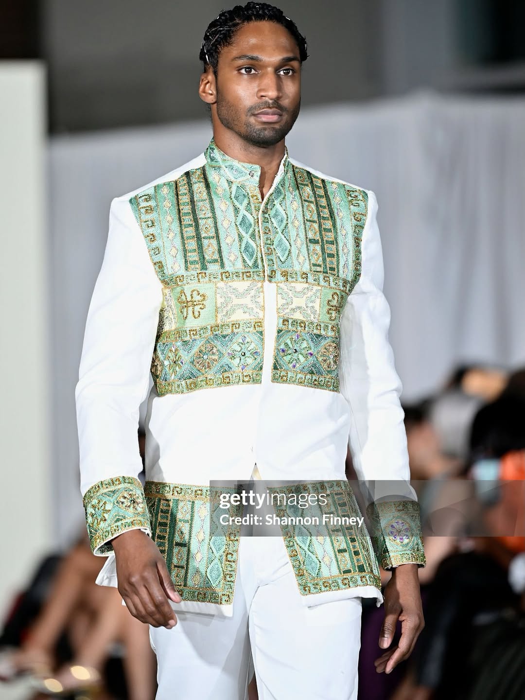 Model wearing a traditional outfit with a green patterned vest on a runway.