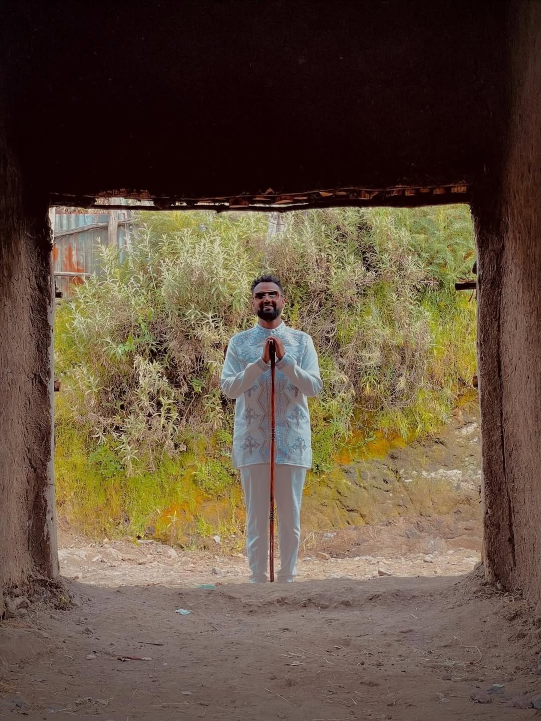Man standing in a natural setting with greenery and a rustic structure. Silver Modern Habesha Couple Outfit Matching Ethiopian Attire ethgebya gebeya usa habesha kemis