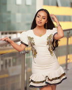 Woman wearing a white dress with gold embroidery standing against a blurred background Modern Ethiopian Couple Set Mini Dress & Men’s Habesha Shirt ethgebya gebeya usa habesha kemis mini