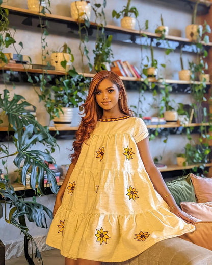 Woman in a yellow dress with floral patterns standing in a room with plants and shelves. Lightweight Short Habesha Dress Modern Summer Ethiopian Kemis ethgebya gebeya habesha kemis usa