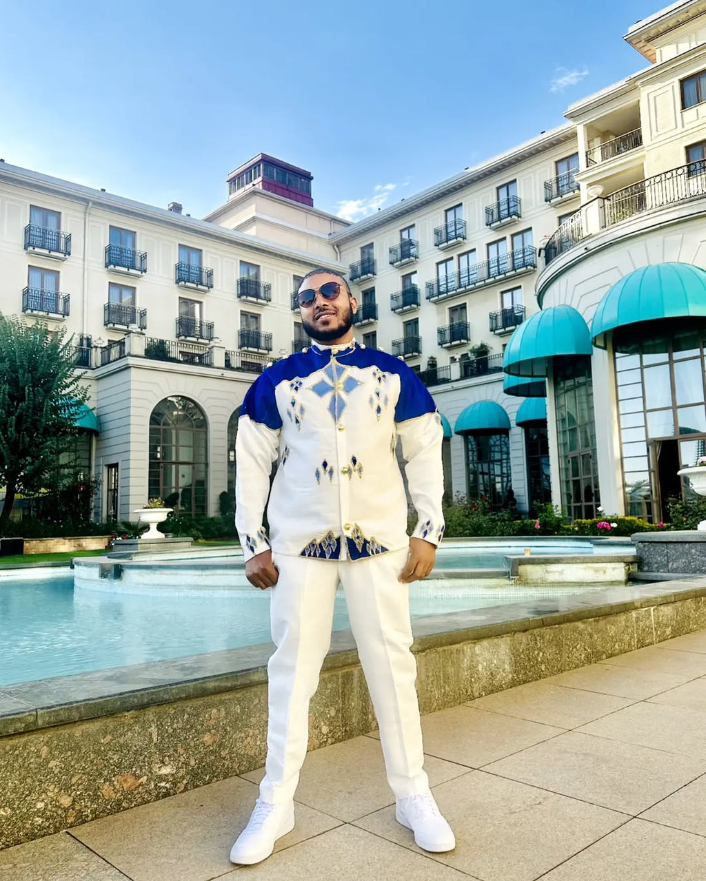 Man in a stylish outfit standing in front of a building with a pool. Blue Habesha Men’s Shirt New Modern Ethiopian Traditional Wear ethgebya gebeya usa habesha mens