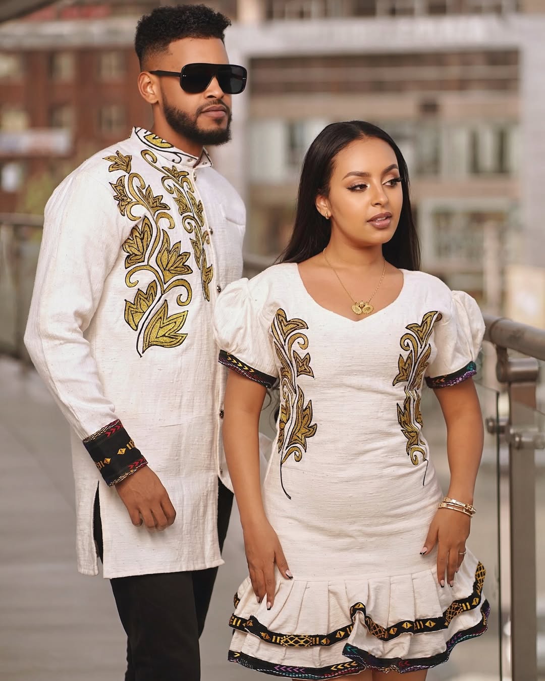 Man and woman in traditional embroidered outfits standing outdoors. Modern Ethiopian Couple Set Mini Dress & Men’s Habesha Shirt ethgebya gebeya usa habesha kemis mini