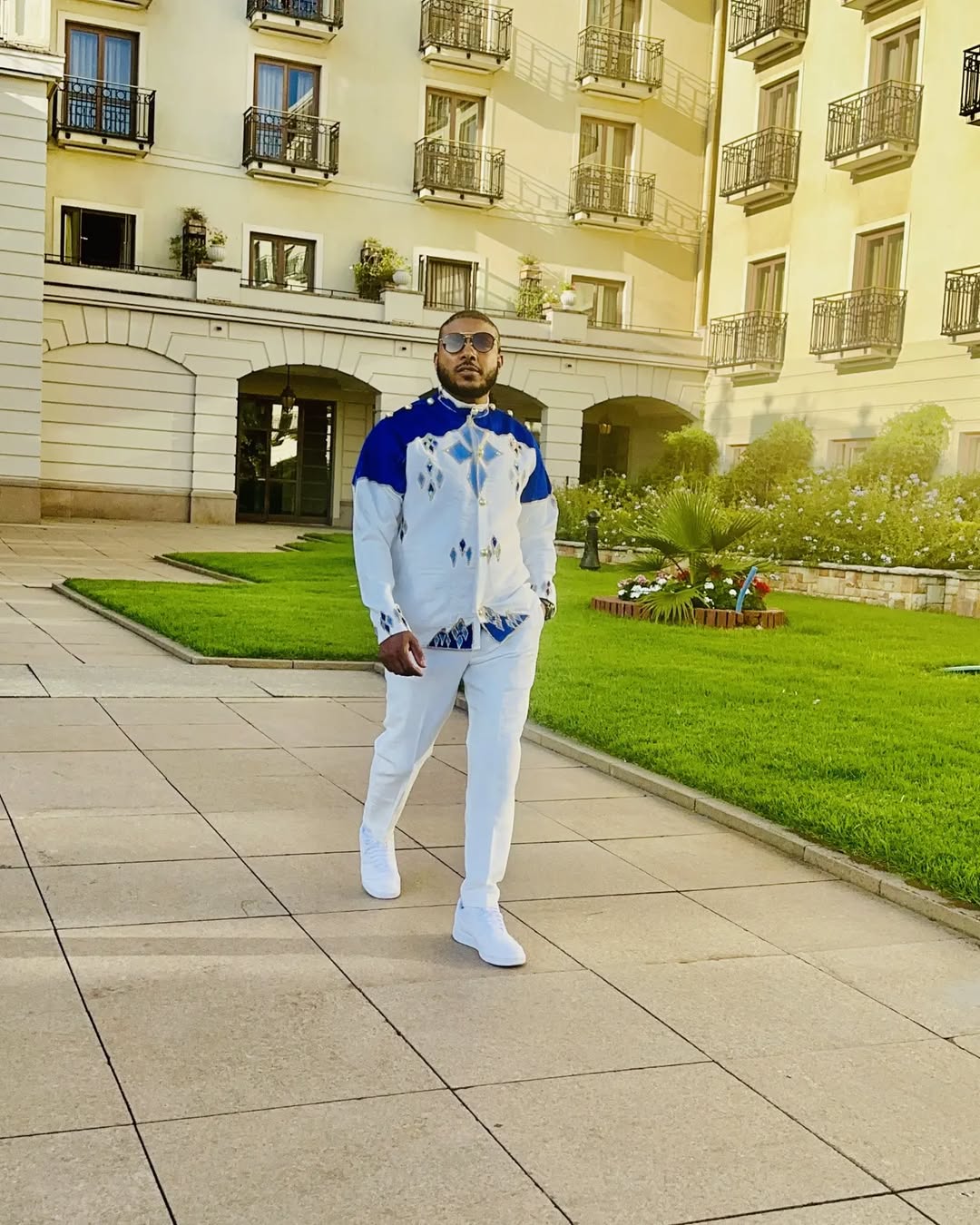 Man in a white outfit with blue accents standing in front of a building with balconies. Blue Habesha Men’s Shirt New Modern Ethiopian Traditional Wear ethgebya gebeya usa habesha mens
