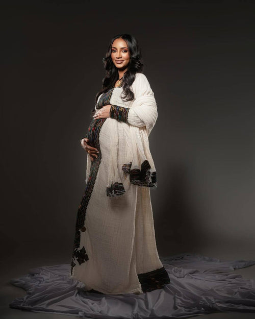 Pregnant woman in a long, light-colored dress with dark accents against a dark background Axum Habesha Kemis & Men’s White Shirt with Gabi Ethiopian Couple Set ethgebya gebeya usa habesha kemis men 
