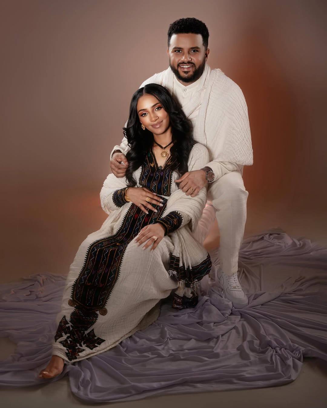 Couple posing together on a purple fabric against a brown background. Axum Habesha Kemis & Men’s White Shirt with Gabi Ethiopian Couple Set ethgebya gebeya usa habesha kemis men 