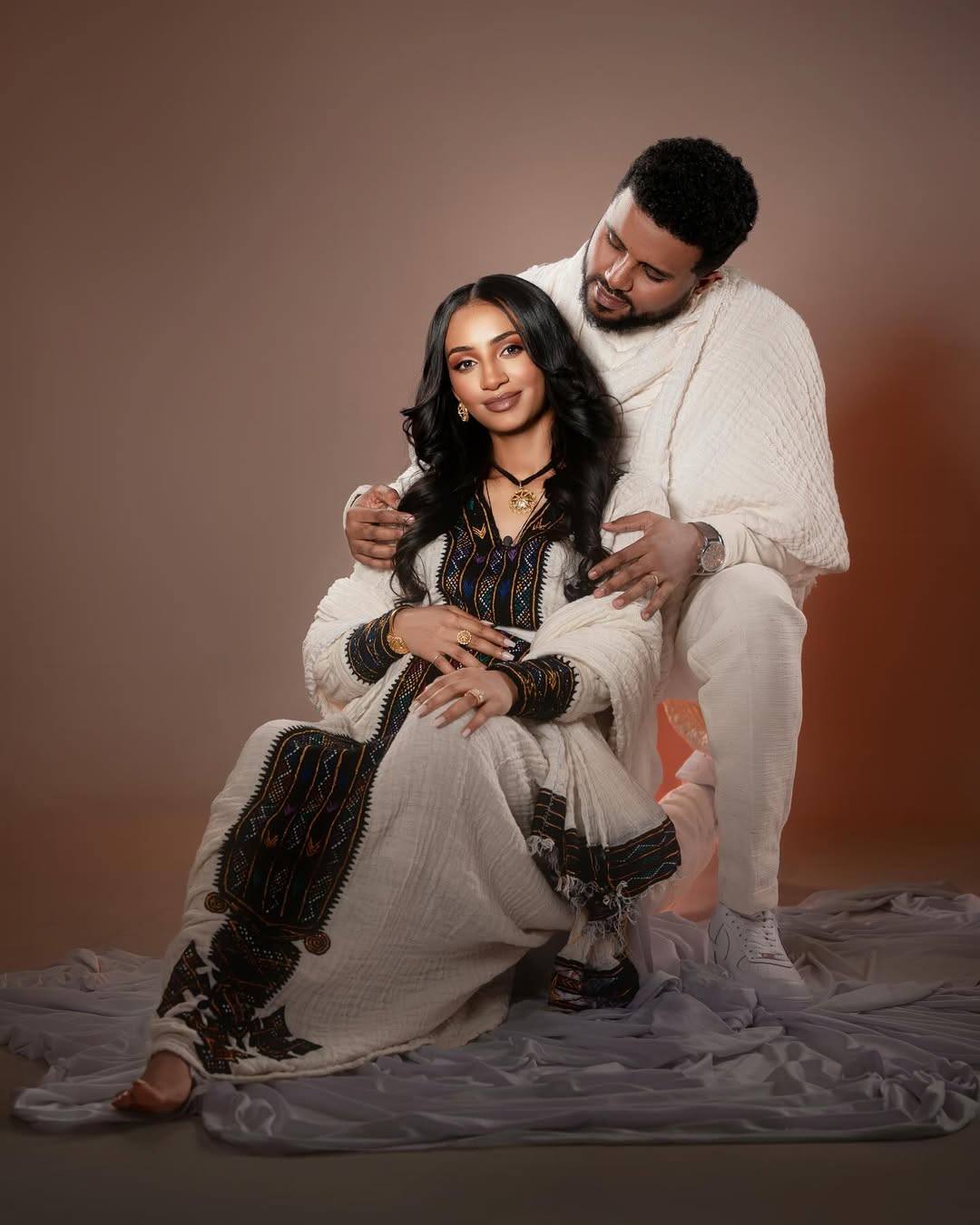Man and woman in traditional attire sitting together on a textured surface with a brown background Axum Habesha Kemis & Men’s White Shirt with Gabi Ethiopian Couple Set ethgebya gebeya usa habesha kemis men 