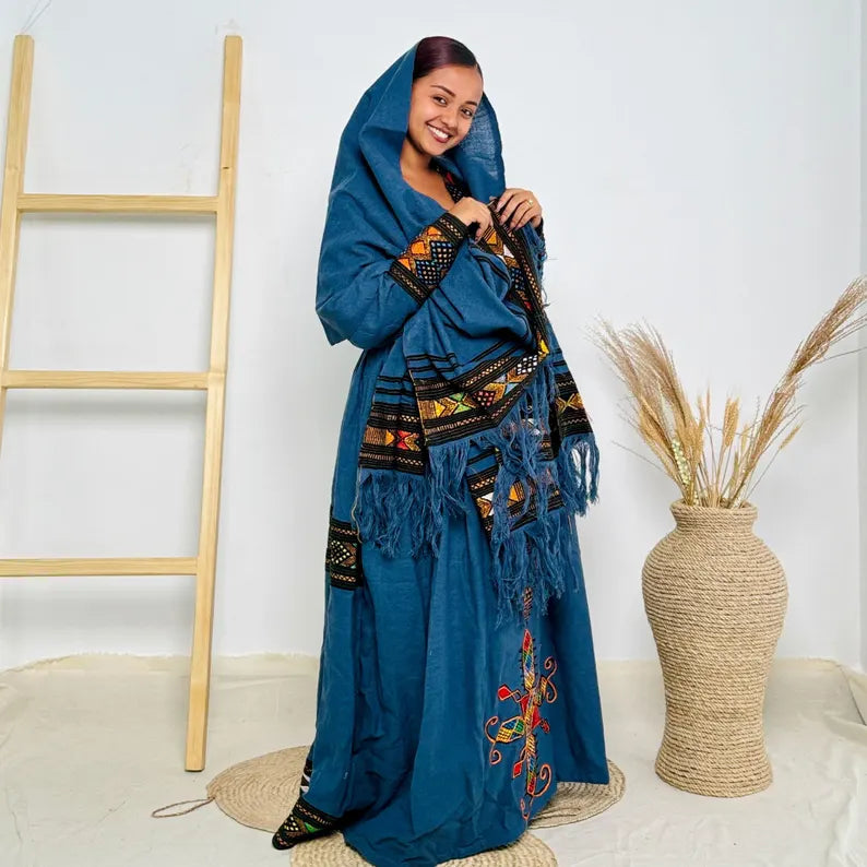 Woman wearing a blue embroidered traditional outfit standing next to a woven vase and wooden ladder. Dark Blue Raya Cultural Dress Handwoven Ethiopian Habesha Kemis ethgebya gebeya usa habesha kemis