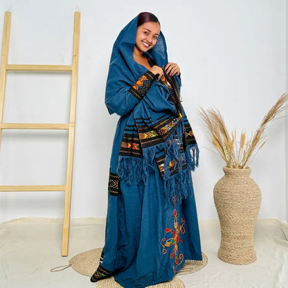 Woman wearing a blue embroidered traditional outfit standing next to a woven vase and wooden ladder. Dark Blue Raya Cultural Dress Handwoven Ethiopian Habesha Kemis ethgebya gebeya usa habesha kemis