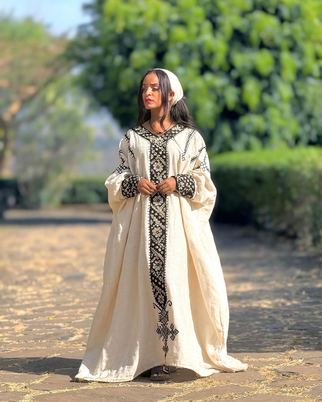 Woman in a long, embroidered cream dress standing outdoors with greenery in the background Gonder Habesha Kemis New Relaxed Ethiopian Traditional Dress ethgebya gebeya usa gondar habesha kemis