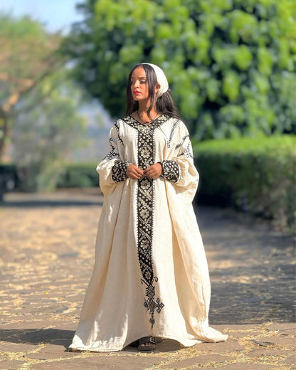 Woman in a long, embroidered cream dress standing outdoors with greenery in the background Gonder Habesha Kemis New Relaxed Ethiopian Traditional Dress ethgebya gebeya usa gondar habesha kemis