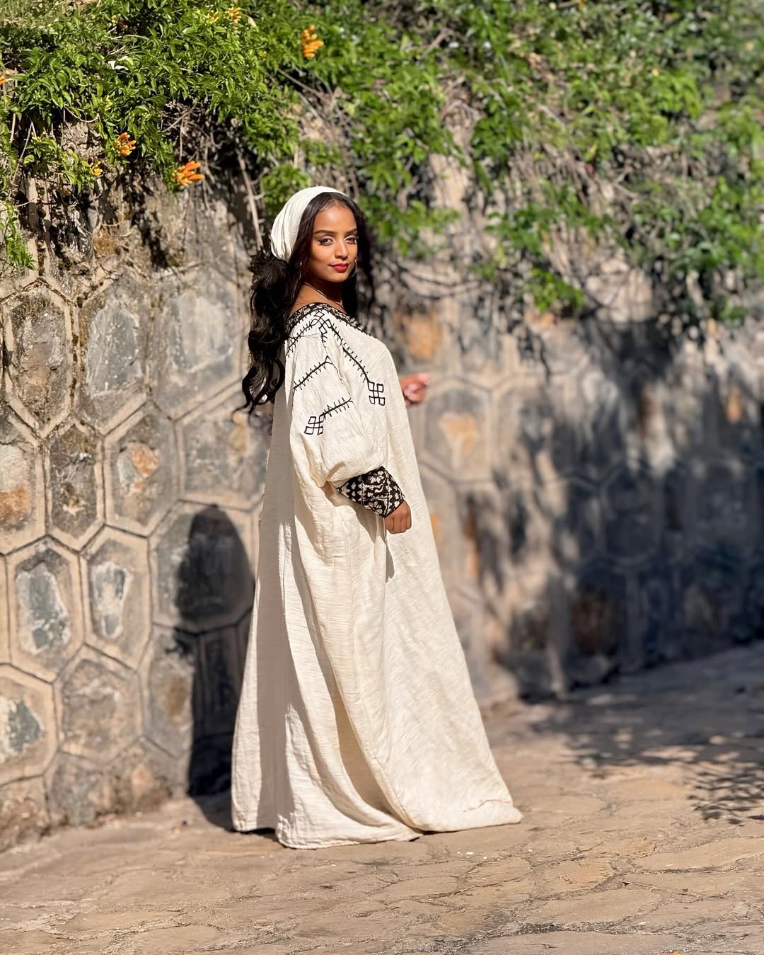 Woman in a long white dress standing against a stone wall with greenery. Gonder Habesha Kemis New Relaxed Ethiopian Traditional Dress ethgebya gebeya usa gondar habesha kemis