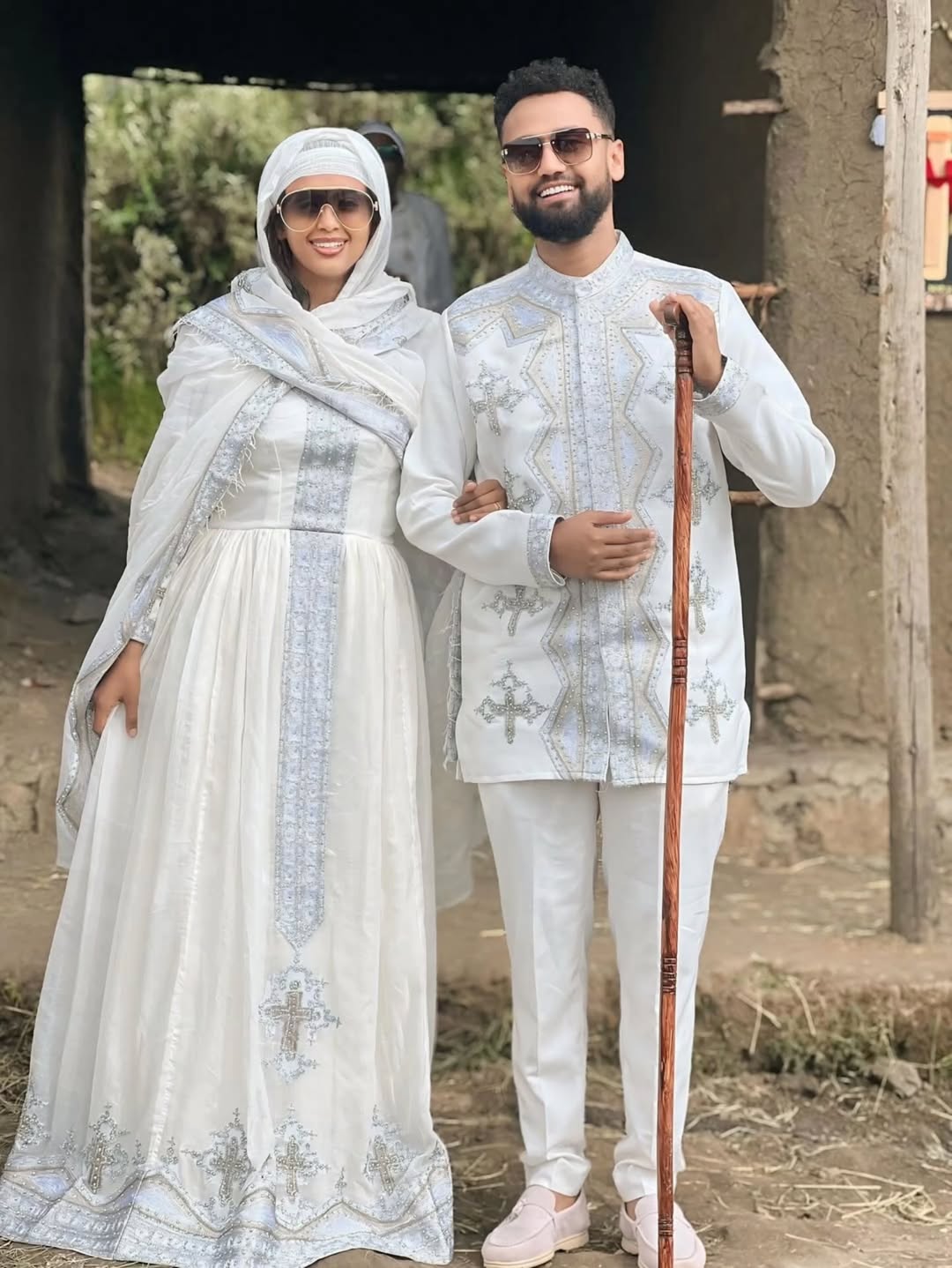 Man and woman in traditional white attire standing outdoors with a rustic background. Silver Modern Habesha Couple Outfit Matching Ethiopian Attire ethgebya gebeya usa habesha kemis