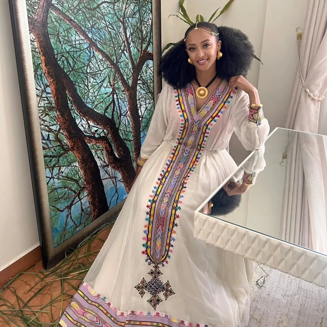 Woman in a white dress with colorful embroidery standing in front of a mirror. Rose Gold Zuria Habesha Kemis Modern Ethiopian Traditional Dress ethgebya gebeya usa habesha kemis
