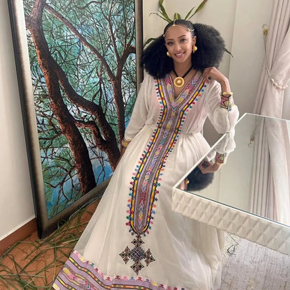 Woman in a white dress with colorful embroidery standing in front of a mirror. Rose Gold Zuria Habesha Kemis Modern Ethiopian Traditional Dress ethgebya gebeya usa habesha kemis