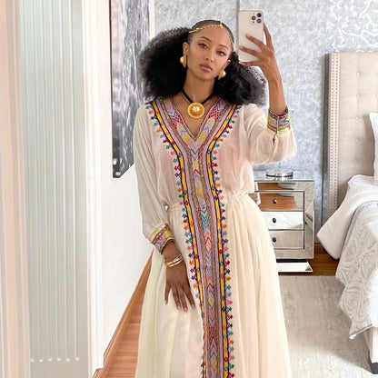 Woman taking a mirror selfie wearing a long, light-colored dress with colorful embroidery in a bedroom. Rose Gold Zuria Habesha Kemis Modern Ethiopian Traditional Dress ethgebya gebeya usa habesha kemis