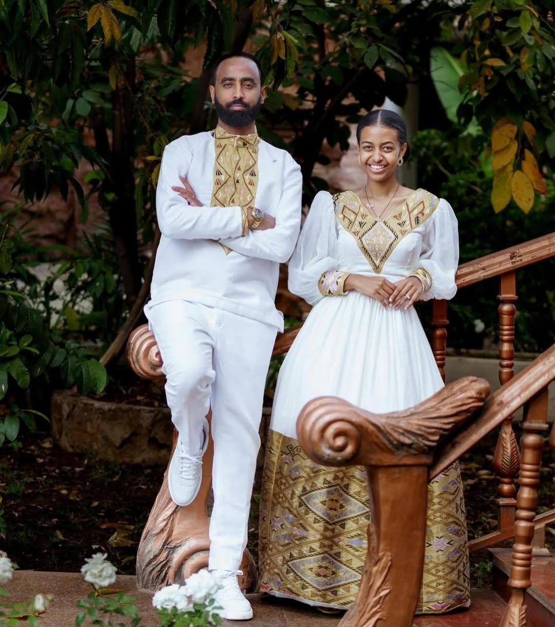 Two people in traditional attire standing outdoors with greenery in the background