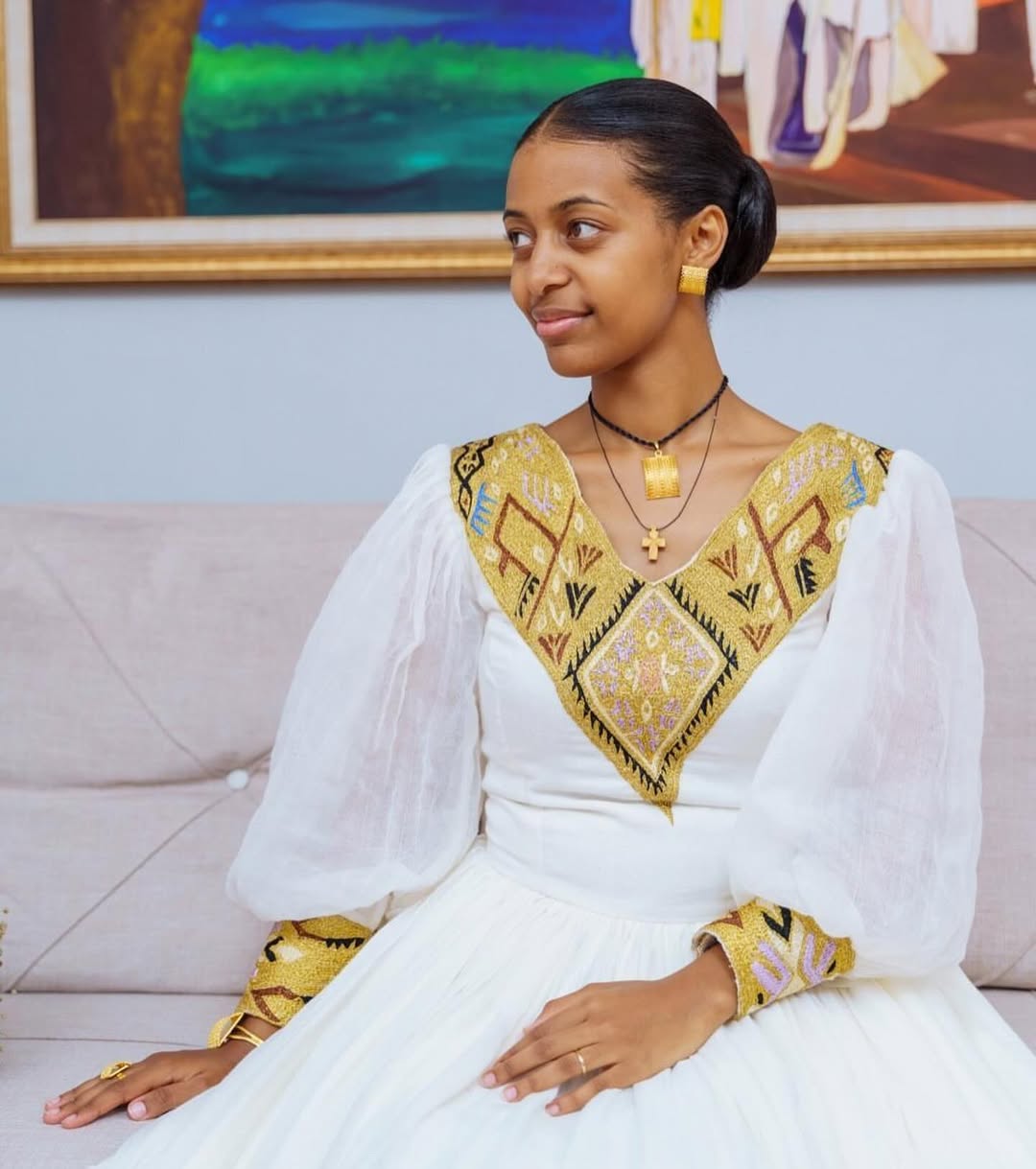 Woman in a white and gold traditional outfit sitting on a couch with a colorful painting in the background.
