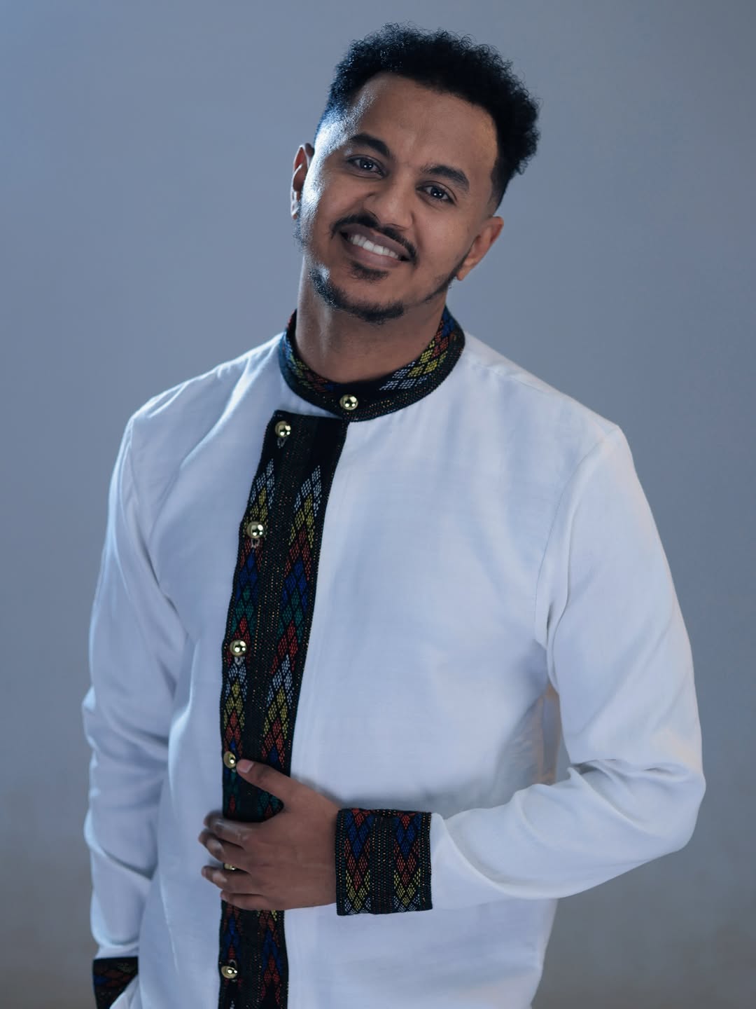 Man wearing a traditional outfit with a white shirt and colorful embroidery on a gray background Ethiopian Men’s Shirt | Handwoven Tilf Embroidery New Habesha Fashion