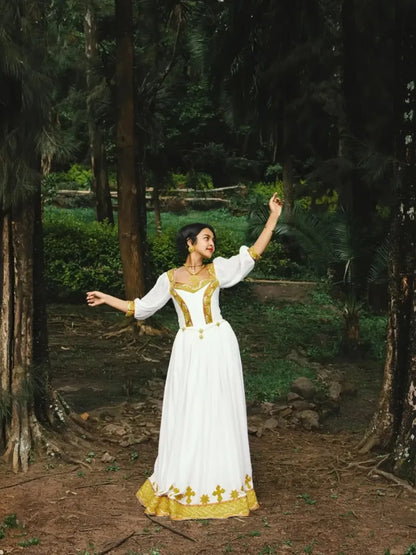 Woman in a white and gold dress standing in a forest. Gold Zuria Habesha Kemis Modern Ethiopian Traditional Dress ethgebya gebeya usa habesha kemis