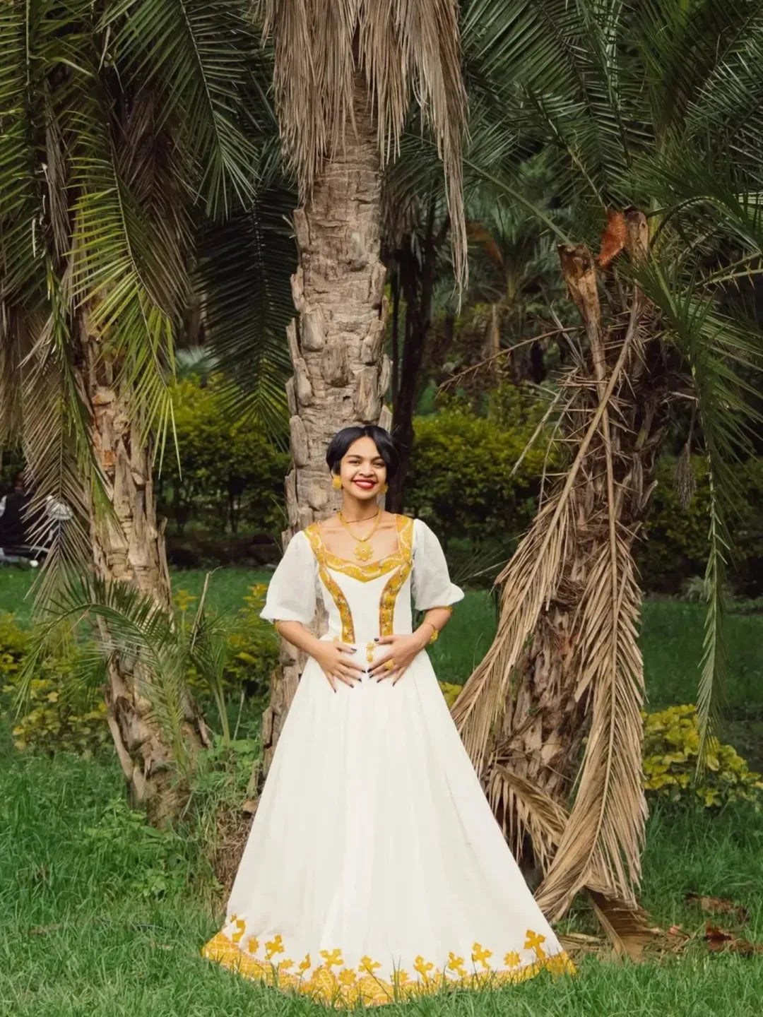 Woman in a white and yellow dress standing in a garden with palm trees. Gold Zuria Habesha Kemis Modern Ethiopian Traditional Dress ethgebya gebeya usa habesha kemis