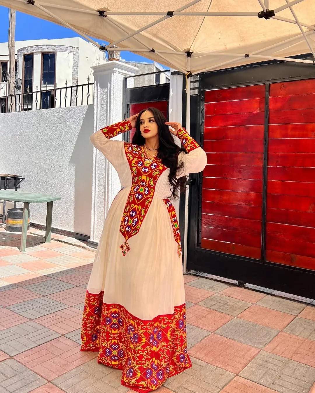 Woman in a colorful traditional outfit standing outdoors under a canopy. Stunning Red Zuria Habesha Kemis Handcrafted Ethiopian Dress  Ethiopian artisanal dress, Habesha style, Habesha fashion, Ethiopian cultural wear, Eritrean cultural attire, Habesha modern outfit, Ethiopian classic dress, Ethiopian embroidery fashion, Habesha traditional clothing, Ethiopian traditional outfits for women, Habesha chic dress, Ethiopian ethnic dress, Habesha inspired fashion, Ethiopian custom made dress, modern Kemiethgbeya