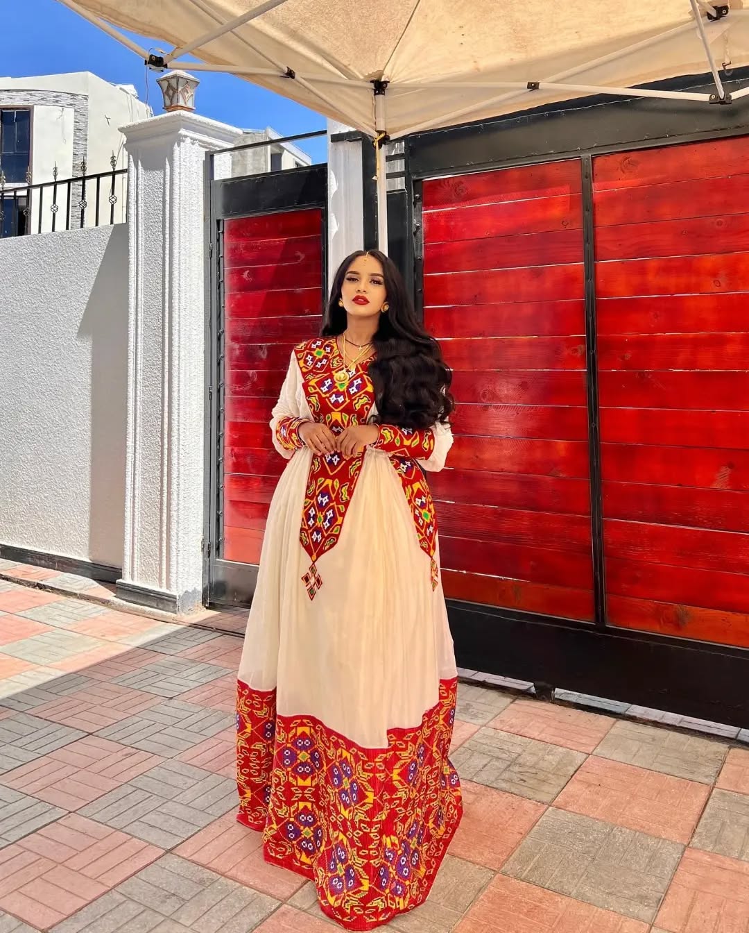 Woman in a traditional outfit standing in front of a red gate. Stunning Red Zuria Habesha Kemis Handcrafted Ethiopian Dress ethgbeya Ethiopian artisanal dress, Habesha style, Habesha fashion, Ethiopian cultural wear, Eritrean cultural attire, Habesha modern outfit, Ethiopian classic dress, Ethiopian embroidery fashion, Habesha traditional clothing, Ethiopian traditional outfits for women, Habesha chic dress, Ethiopian ethnic dress, Habesha inspired fashion, Ethiopian custom made dress, modern Kemis, Ethiopi