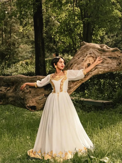 Woman in a white dress with gold accents standing next to a large tree trunk in a forest setting. Gold Zuria Habesha Kemis Modern Ethiopian Traditional Dress ethgebya gebeya usa habesha kemis