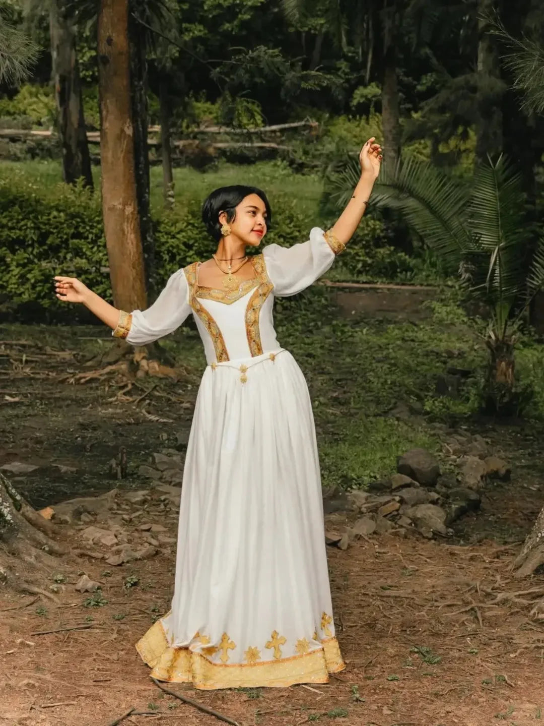 Woman in a white and gold dress standing in a natural setting with trees and greenery. Gold Zuria Habesha Kemis Modern Ethiopian Traditional Dress ethgebya gebeya usa habesha kemis