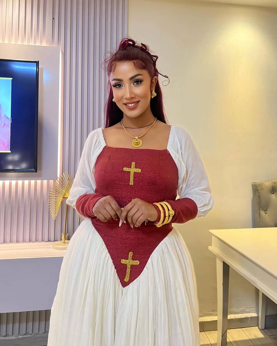 Woman wearing a red and white outfit with gold crosses indoors. Elegant  Modern Red-Accent Habesha Kemis Ethgebya