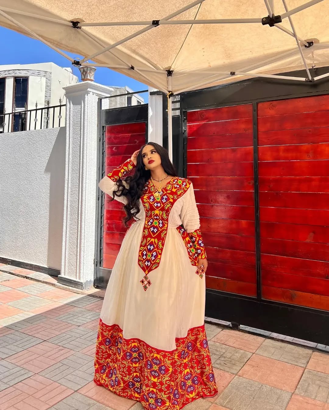 Woman in a traditional outfit with red and gold patterns standing outdoors. Stunning Red Zuria Habesha Kemis Handcrafted Ethiopian Dress  Ethiopian artisanal dress, Habesha style, Habesha fashion, Ethiopian cultural wear, Eritrean cultural attire, Habesha modern outfit, Ethiopian classic dress, Ethiopian embroidery fashion, Habesha traditional clothing, Ethiopian traditional outfits for women, Habesha chic dress, Ethiopian ethnic dress, Habesha inspired fashion, Ethiopian custom made dress, modern Kethgbeya