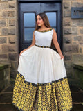 Woman wearing a white top with black and gold accents and a yellow floral skirt in front of a stone building.