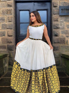 Woman wearing a white top with black and gold accents and a yellow floral skirt in front of a stone building.