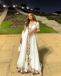 Woman in a white dress with gold and purple details standing on a stone path at night.