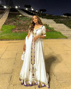 Woman in a white dress with gold and purple details standing on a stone path at night.