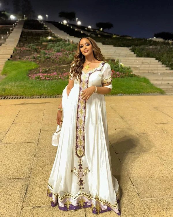 Woman in a white dress with gold and purple details standing on a stone path at night.