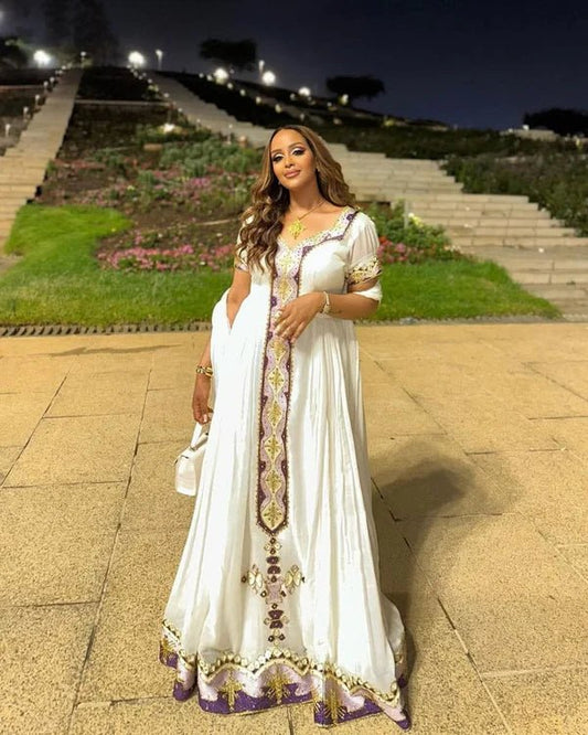 Woman in a white dress with gold and purple details standing on a stone path at night.
