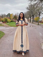 Woman in a long, beige dress with blue embroidery standing on a street. Beige Modern Zuria Habesha Kemis Stylish Ethiopian Traditional Dress ethgebya gebeya habesha kemis