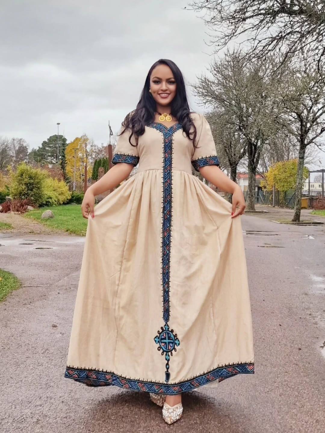 Woman wearing a long, beige dress with blue embroidery standing on a street. Beige Modern Zuria Habesha Kemis Stylish Ethiopian Traditional Dress ethgebya gebeya habesha kemis