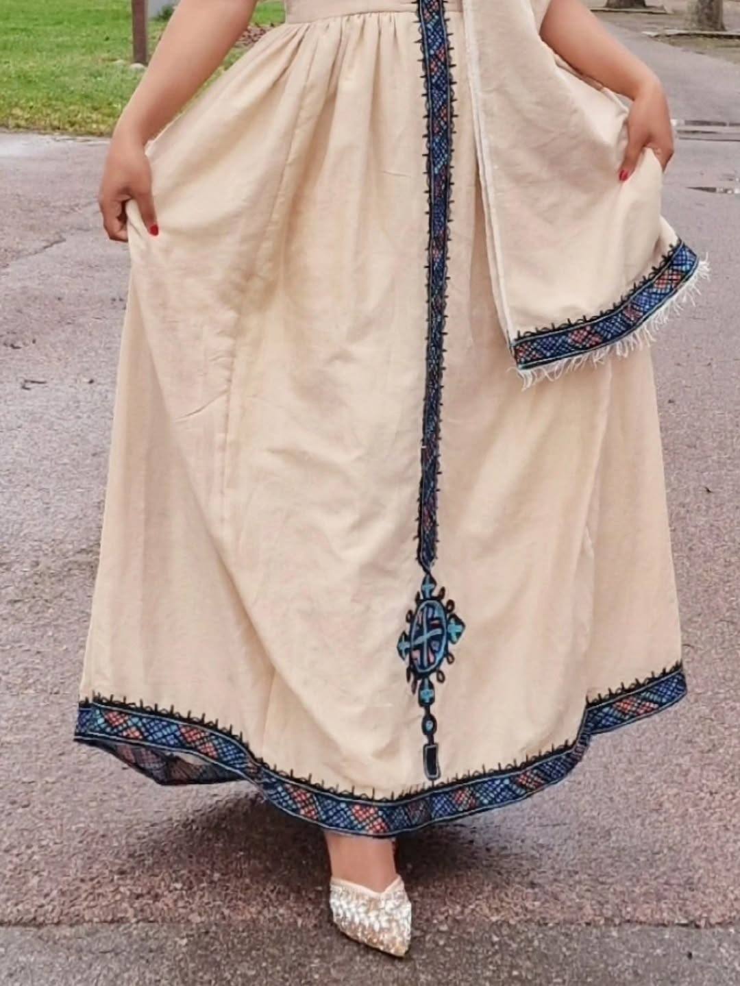 Woman wearing a traditional outfit with a beige dress and blue embroidery on a road. Beige Modern Zuria Habesha Kemis Stylish Ethiopian Traditional Dress ethgebya gebeya habesha kemis