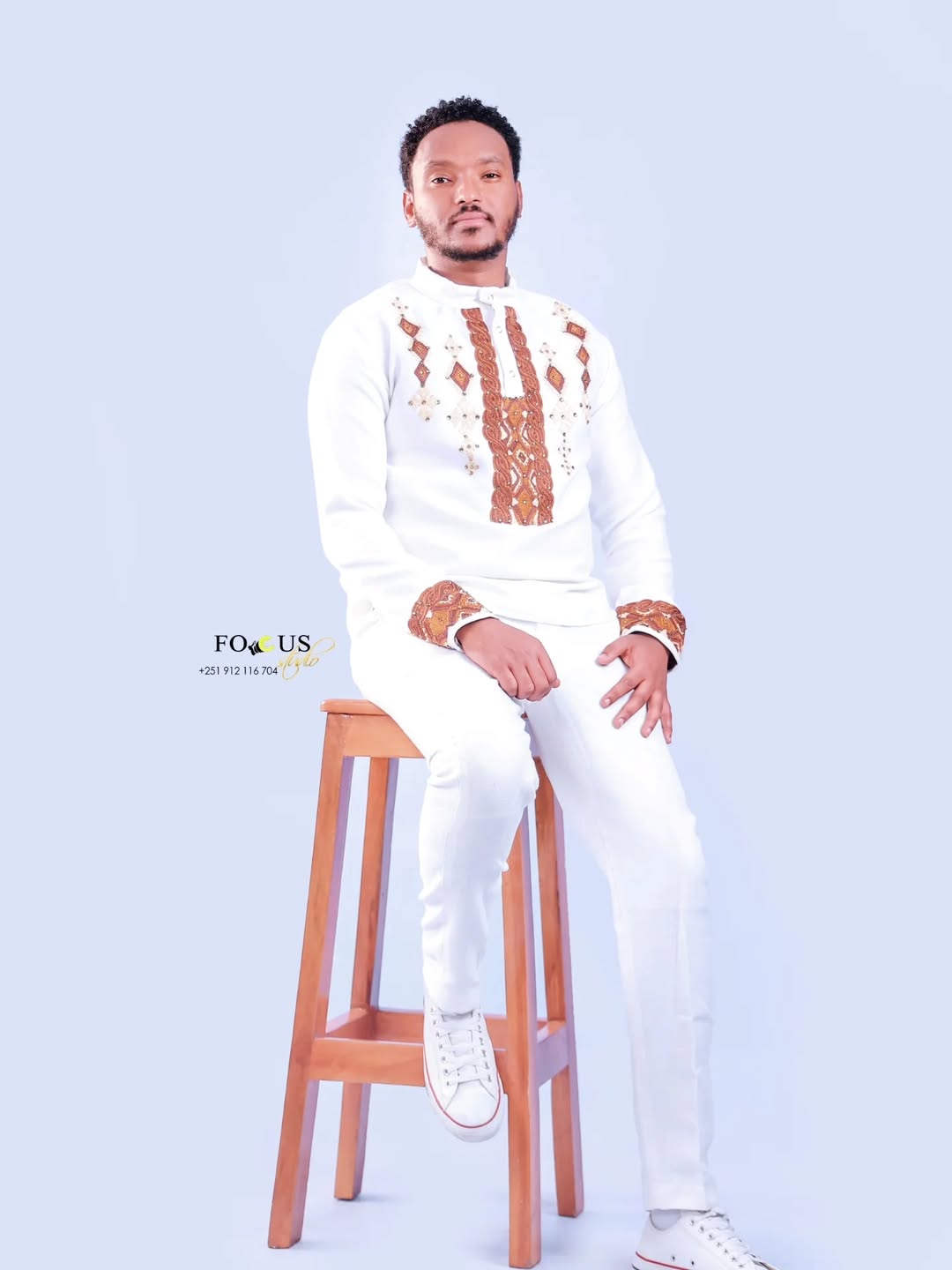 Man in a white outfit with brown patterns sitting on a wooden stool against a light blue background. Ethiopian gown, Habesha wedding dress, Eritrean evening dress, Ethiopian traditional gown, Ethiopian heritage dress, Habesha chic, Eritrean cultural attire, Habesha elegant dress, Eritrean bridal wear, Ethiopian vintage dress, Habesha bridesmaid dress, African ethnic dress, Ethiopian handcrafted dress, Eritrean handcrafted dress, traditional African gown, Habesha festive dress, Ethiopian formal wear,