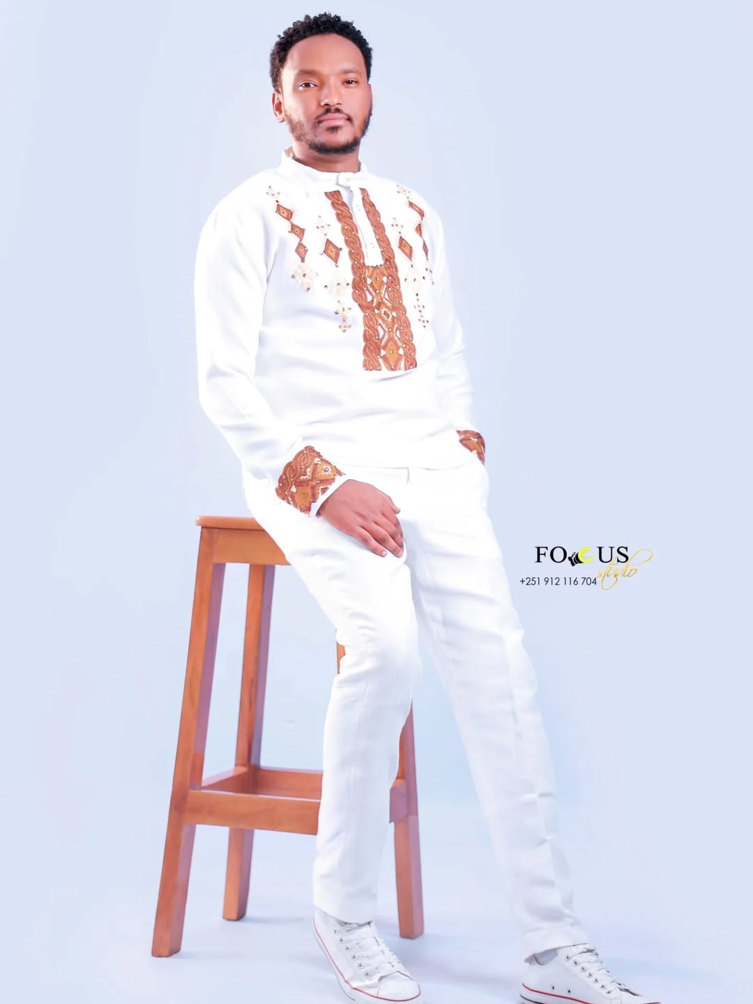 Man in a white outfit with brown patterns standing next to a wooden stool on a light blue background. Ethiopian gown, Habesha wedding dress, Eritrean evening dress, Ethiopian traditional gown, Ethiopian heritage dress, Habesha chic, Eritrean cultural attire, Habesha elegant dress, Eritrean bridal wear, Ethiopian vintage dress, Habesha bridesmaid dress, African ethnic dress, Ethiopian handcrafted dress, Eritrean handcrafted dress, traditional African gown, Habesha festive dress, Ethiopian formal wear,