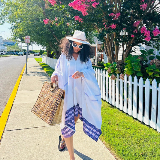Woman in a white outfit with a blue pattern, white hat, and sunglasses walking on a sidewalk with a white picket fence and pink flowers in the background. modern ethiopian dress habesha traditional dress habesha dress online shopping ethiopian habesha kemis ethiopian kemis habesha libs ethiopian new dress habesha attire fashion ethiopian dress ethiopian women's dress women's ethiopian clothing modern ethiopian traditional clothes habesha cultural dress habesha traditional cloth new ethiopian traditional dre