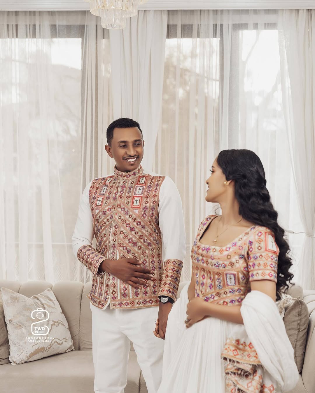 Couple in traditional attire standing in a well-lit room with white curtains. Ethiopian Brown Habesha Wedding Attire Matching Couple Set Ethgebya
